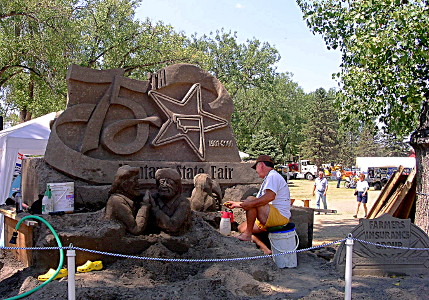 [Part of the sculpture is complete and has a big 75 and a star with the shape of Montana in it. A ribbon below that says Montana State Fair. A man is working on the sculpture to add people in front of it.]